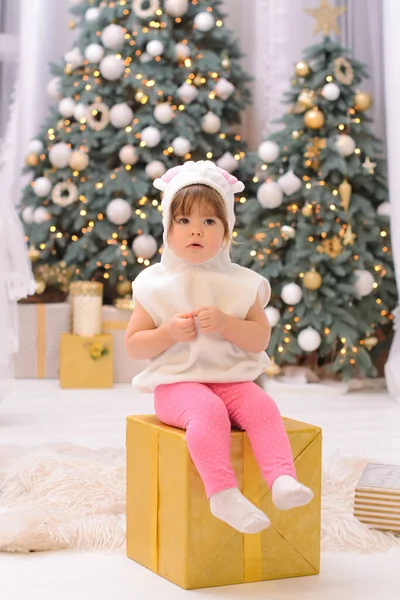 一个身穿白色兔子衣服的小女孩坐在一个巨大的金色礼品盒上 靠着圣诞树的背景 — 图库照片