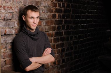 Siyah ceketli bir gencin portresi kahverengi, kırmızı ve siyah tuğlalı bir duvarın arkasında.