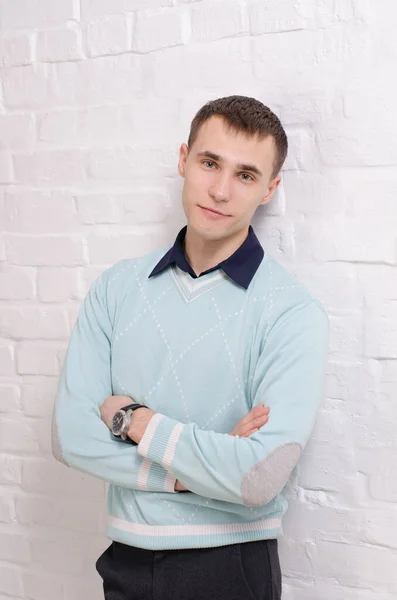 一个穿着蓝色套头衫和黑色裤子的年轻人站在一面白色的砖墙旁边 他的画像交叉在他的胳膊上 — 图库照片