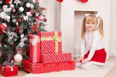 Beyaz elbiseli ve kırmızı Noel Baba etekli bir kız, Noel ağacının yanında oturuyor ve hediyelerle dolu kutular, dekoratif bir şöminenin arka planında, parlak bir odada.