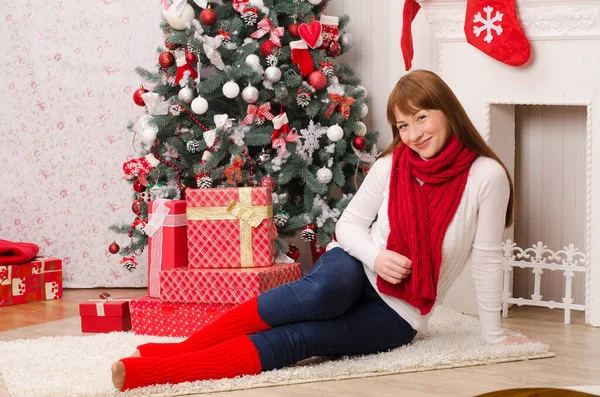 一个身穿白色毛衣 头戴红色围巾 头戴高尔夫球的长发女孩 坐在圣诞树旁 在明亮房间的壁炉前 拿着礼物盒 — 图库照片