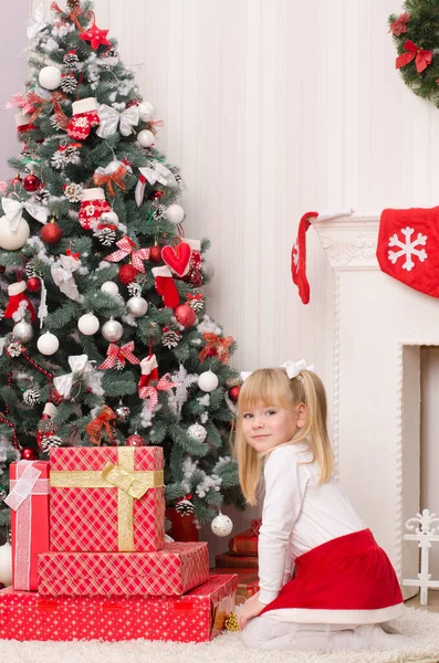 白い服を着た女の子と赤いサンタのスカートは 明るい部屋で クリスマスツリーとギフト付きの箱の近くに座っています — ストック写真