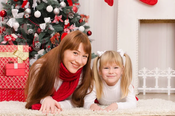 快乐的母亲和女儿躺在圣诞树旁的地毯上 戴着礼物的红盒子 背靠明亮房间里的壁炉 — 图库照片