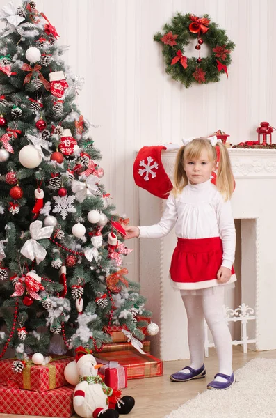 白い服の少女と赤いサンタのスカートは 明るい部屋で 装飾的な暖炉の背景に クリスマスツリーの近くに立っています — ストック写真
