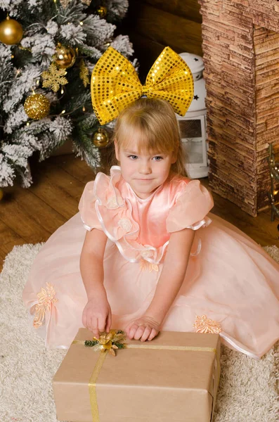 身着节日舞会礼服 头上顶着巨大黄色蝴蝶结的女孩 手里拿着圣诞礼物 坐在地板上的地毯上 — 图库照片