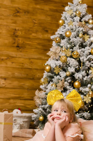 頭に大きな黄色い弓を持つ少女は木の壁に囲まれた部屋の床に横たわっていますクリスマスツリーを背景に — ストック写真