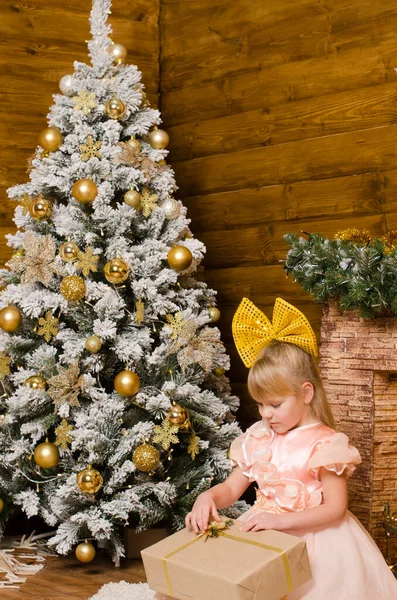 クリスマスツリーと暖炉の背景を背景に 木製の壁を持つ部屋の床にラグの上に座って 彼女の頭の上に巨大な黄色の弓を持つお祝いのボールのガウンで幸せな女の子 ギフトボックスをアンパックする — ストック写真