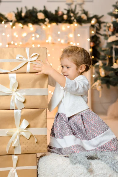 一个漂亮而快乐的女孩坐在地板上 靠着一个有花环灯的圣诞壁炉 把装有礼物的盒子堆在一起 — 图库照片