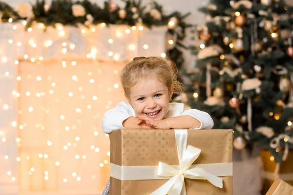 美丽而快乐的女孩坐在地板上 手里拿着大的金色礼品盒 上面系着白色的缎带 在新年的背景下 壁炉和圣诞树层出不穷 — 图库照片