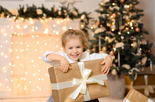 美丽而快乐的女孩坐在地板上 手里拿着大的金色礼品盒 上面系着白色的缎带 在新年的背景下 壁炉和圣诞树层出不穷 — 图库照片