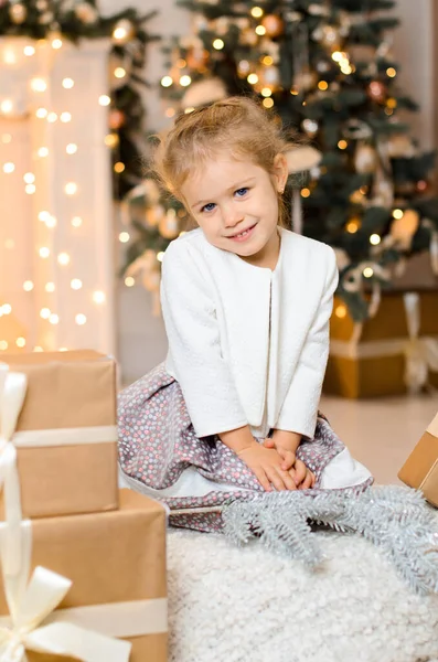 白いセーターを着た美しい少女がクリスマスツリーを背景にリボン付きのギフトボックスの近くに座って暖炉の上に輝くガーランド ボケライト — ストック写真