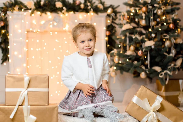 一个穿着白色毛衣的漂亮姑娘坐在礼品盒旁 背靠着一棵圣诞树 壁炉上挂着一个闪闪发光的花环 灯火通明 — 图库照片
