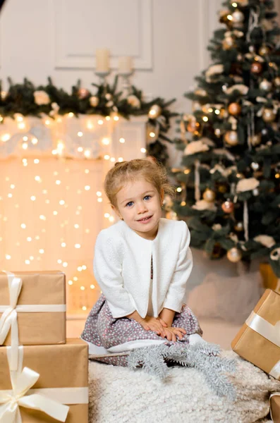 一个穿着白色毛衣的漂亮姑娘坐在礼品盒旁 背靠着一棵圣诞树 壁炉上挂着一个闪闪发光的花环 灯火通明 — 图库照片
