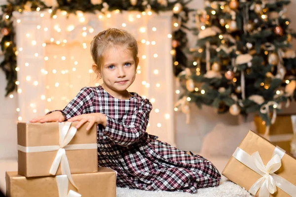 一个身穿格子花裙的漂亮姑娘坐在礼品盒旁 背靠着一棵圣诞树和一个闪闪发亮的花环 灯火通明 — 图库照片