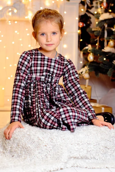 一个穿着格子呢衣服的漂亮姑娘坐在圣诞树的后面 一个闪闪发亮的花环和吊灯旁边 — 图库照片