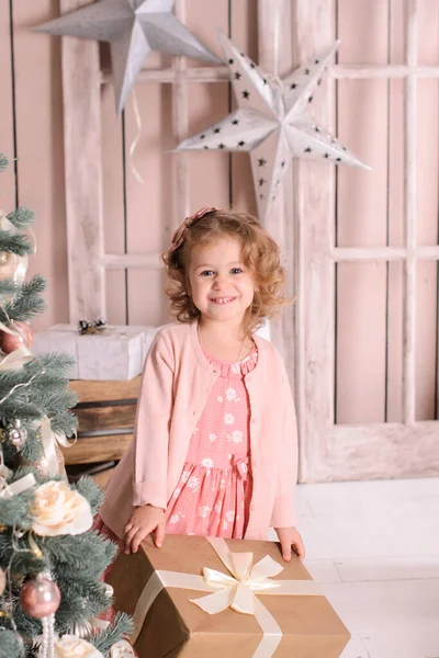 一个穿着粉色衣服的美丽的微笑的女孩从一棵装饰着球的圣诞树后面向外张望 手里拿着一个礼品盒 — 图库照片