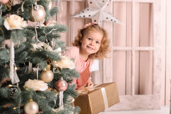 一个穿着粉色衣服的美丽的微笑的女孩从一棵装饰着球的圣诞树后面向外张望 手里拿着一个礼品盒 — 图库照片