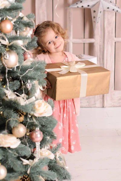 ピンクの服を着た美しい笑顔の女の子が玉で飾られたクリスマスツリーの後ろからのぞき見彼女の手にギフトボックスを持っています — ストック写真