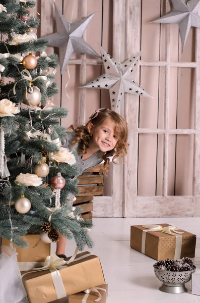 一个美丽的微笑的女孩 从一棵装饰过的圣诞树后面 靠着一堵有星星的阁楼木墙 带着礼物在地板上嬉笑地偷窥着 — 图库照片