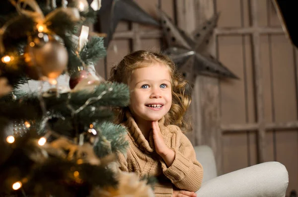 一个穿着米黄色毛衣的漂亮的笑姑娘坐在一把浅灰色的扶手椅上 手托着脸 背靠着一堵有星星和圣诞树的褐色木墙 — 图库照片