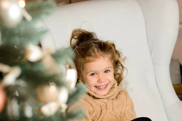 穿着米黄色毛衣的漂亮的笑女孩坐在一棵圣诞树后的一把浅灰色扶手椅上 — 图库照片