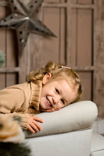 一个穿着米黄色毛衣的漂亮的笑姑娘坐在一把浅灰色的扶手椅上 把头靠在扶手上 背靠着一堵带有星星的褐色木墙 — 图库照片