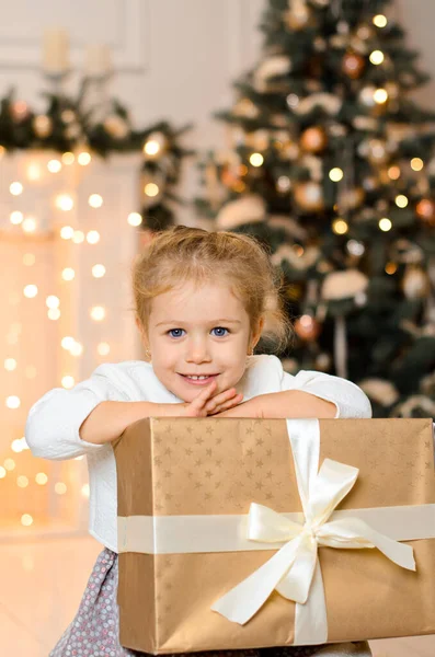 白いリボンで結ばれた大きな黄金のギフトボックスを持って床に座っている美しい幸せな笑いの女の子 ガーランド クリスマスツリーの新年のボケを背景に ロイヤリティフリーのストック写真