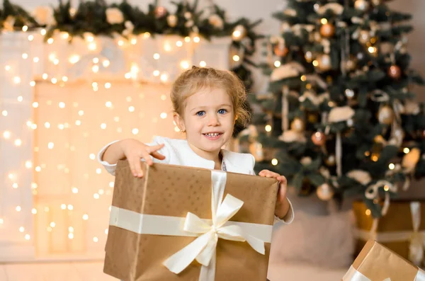 白いリボンで結ばれた大きな黄金のギフトボックスを持って床に座っている美しい幸せな笑いの女の子 ガーランド クリスマスツリーの新年のボケを背景に ストックフォト