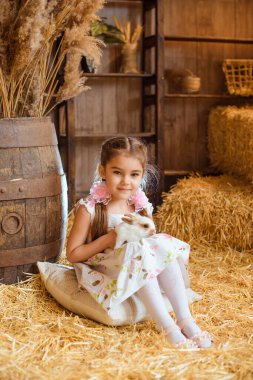 Saçında gül şeklinde iki fiyonklu bir kız beyaz bir yastığa oturur ve arka planda beyaz bir tavşan tutar..