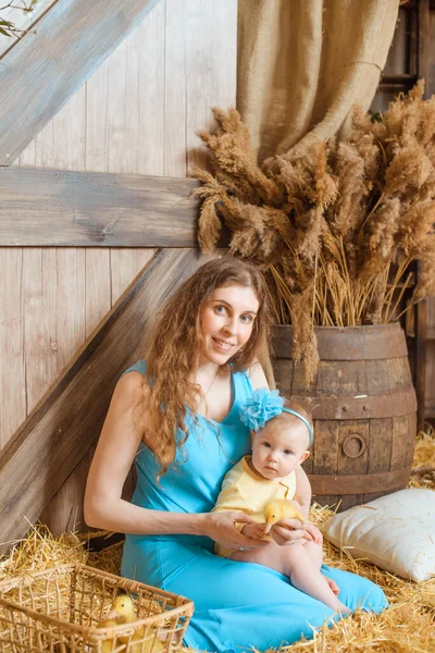 青いドレスを着た波状の髪の美しい女性が腕の中に幼児を抱えている干し草の俵に座っています — ストック写真