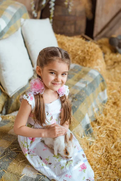 彼女の髪にバラの形をした2つの弓を持つ笑顔の女の子は納屋の背景に白いウサギを保ちます — ストック写真