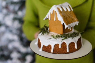 Yeşil elbiseli bir adam, elinde zencefilli kurabiye evi tutuyor, karla kaplı bir Noel ağacının zeminine karşı.