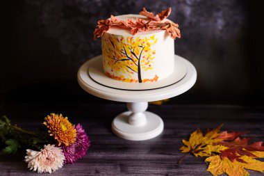 Krizantem yanında sonbahar desenli bir pasta, beyaz bir stantta ve koyu bir arka planda.