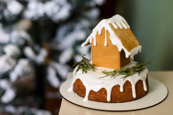 在白冰蛋糕上的姜饼屋 背景是被雪覆盖的圣诞树 — 图库照片