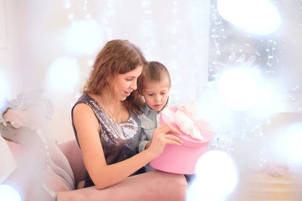 快乐的母亲和儿子坐在椅子上 打开装有礼物的盒子 靠着窗上闪闪发光的花环 朝前看去 — 图库照片