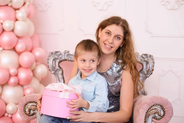 妈妈留着长长的波浪般的头发 怀里抱着她的儿子 还有一个带着白色缎带的粉色礼品盒 — 图库照片