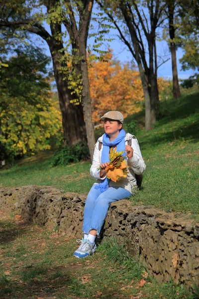 在照片中 一个拿着一束黄叶的年轻女子坐在那里 梦想着秋天公园里的什么 — 图库照片