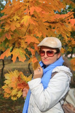 Fotoğraf, güneş gözlüklü, sarı akçaağaç yapraklı genç ve çekici bir kadının portresini gösteriyor..