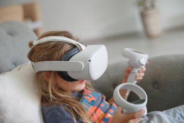 Evde video oyunu oynayan VR maskeli küçük bir çocuğun fotoğrafı. Sanal gerçekliğin simülasyonu. Akıllı çocuk siber alemden hoşlanır. Çevrimiçi rekabet.