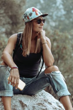 Güzel, neşeli bir kadının portresi. Dağ parkındaki taşın üzerinde dinleniyor. Elinde telefon var. Yaz günü gezintiye çıkmak. Mutlu yaz haftasonları.