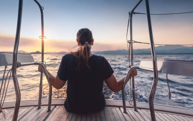 Yelkenlinin güvertesinde oturan ve manzaranın tadını çıkaran bir kadının arka görüntüsü. Hafif gün batımı ışığında deniz. Huzurlu bir yat yolculuğu..