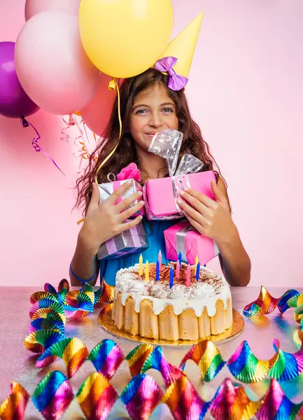 Happy girl with gift boxes in hands enjoying her birthday party. Colorful air balloons, garland and festive cake with candles create a festive mood.