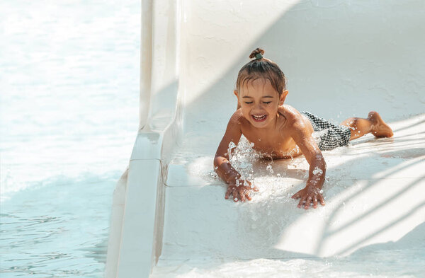 Маленький веселый ребенок мчится вниз на высокой скорости на водной горке в аквапарке в жаркий солнечный день. Счастливый мальчик наслаждается летними каникулами.