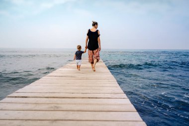Annenin kıymetli oğlunun elini tutarken ve tahta iskelede yürürken görüntüsü. Birlikte harika deniz manzarasının tadını çıkarıyoruz. Aile sevgisi kavramı.