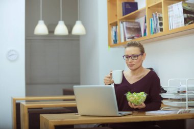 Taze yeşil sebze salatası yiyen ve ofiste kahve içen hoş bir kadının portresi. Uzaktan kumanda. Sağlıklı bir şeyler atıştırmak için mola zamanı..