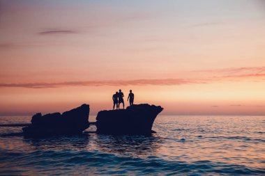 Denizin ortasında kayaların üzerinde duran üç arkadaşın siluetleri. Gün batımında okyanusa atlayan uçurum. Yaz eğlencesi yaşam tarzı.