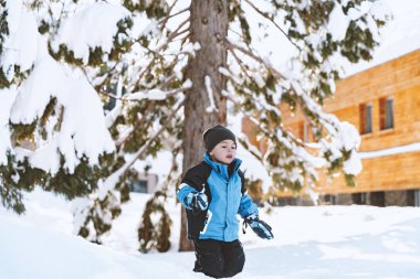 Sıcak ve rahat kış kayak kıyafetleri içinde neşeli bir çocuk, aydınlık bir günde yumuşak karda mutlu bir şekilde oynar. Kış oyunları açık havada