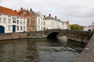 Bruges, Belçika - 13 Eylül 2022: Taş bir köprünün geçtiği kanal boyunca tuğla evler. Burada bulutlu bir günde görebilirsiniz.