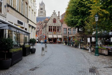 Bruges, Belgium - September 9, 2022: There are houses by the paved street. There is a small square next to it and a bit further you can see the tower of the cathedral, there are a few people there