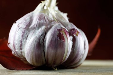 One ripe garlic bulb is placed on a wooden board and one red leaf
